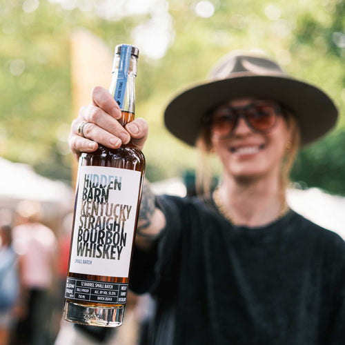 Person holding a bottle of Hidden Barn Kentucky Straight Bourbon Whiskey with a blurred outdoor background