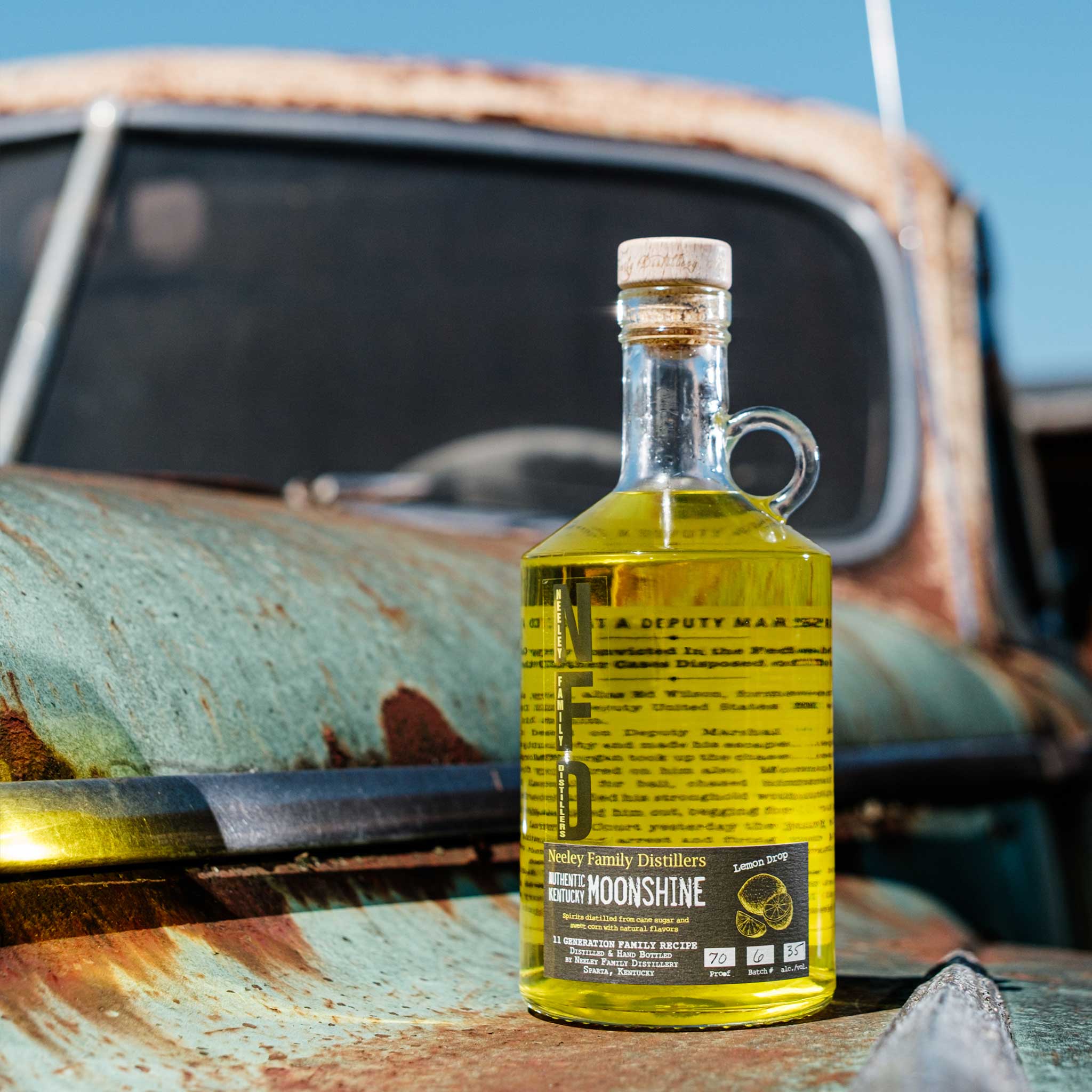 Bottle of Neeley Family Distillery Moonshine on a rusty vehicle with a blue sky background