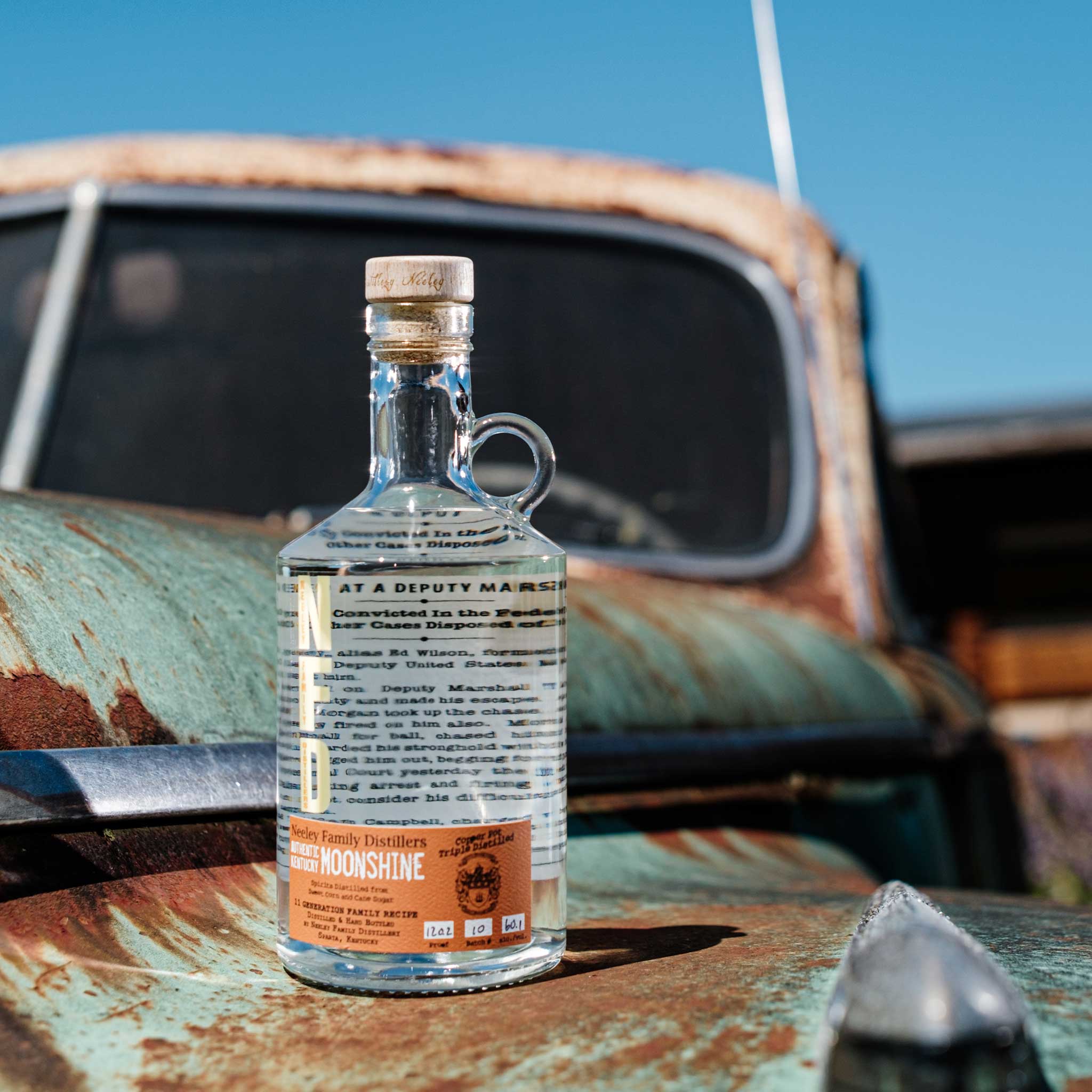 Bottle of Neeley Family Distillery Moonshine on a rusty vehicle with a blue sky background